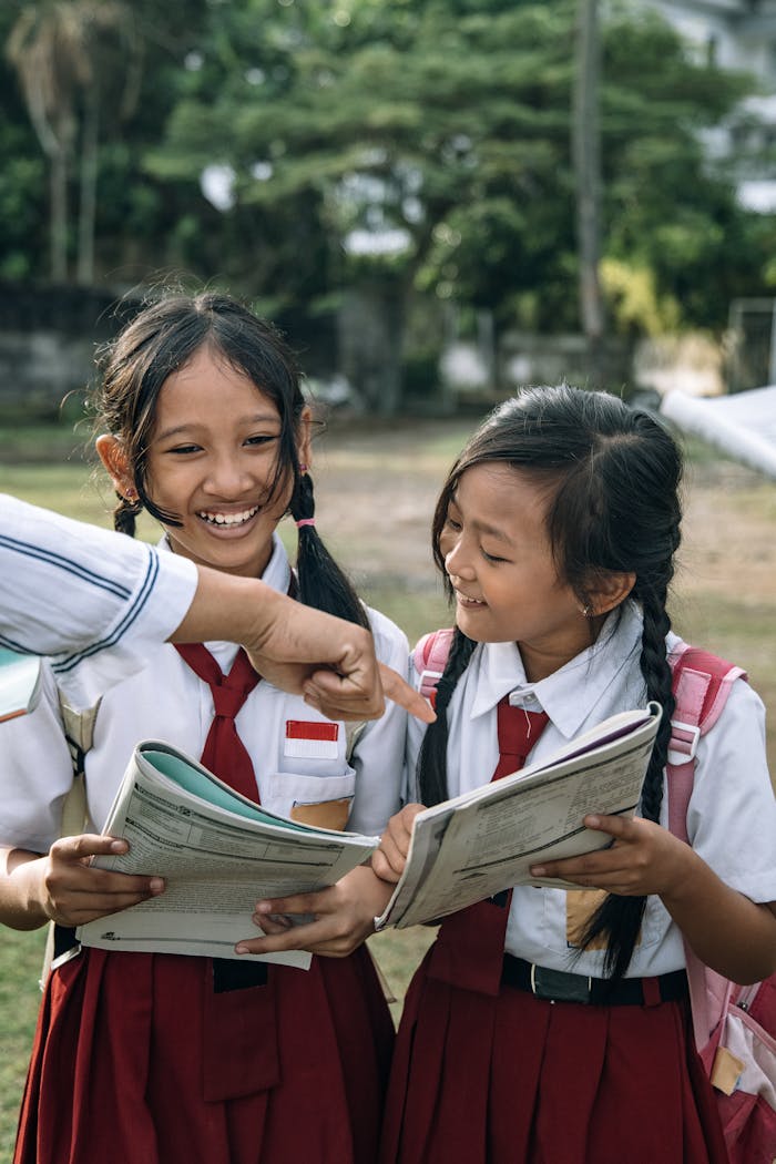 Two schoolgirls in uniforms smiling and reading book together outdoors.